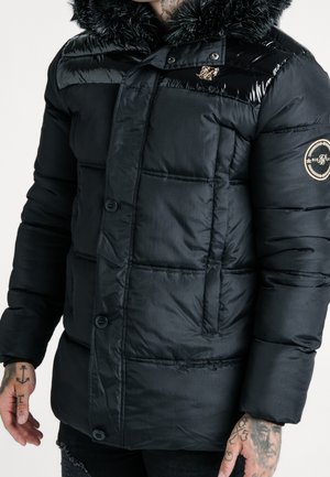 Uomo che indossa una giacca invernale imbottita nera con cappuccio bordato di pelliccia e patch con logo sul petto e sulla manica, mostrando le mani tatuate.