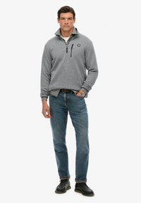 Szary polar z zamkiem przy kołnierzu, w połączeniu z koszulą w kratę, niebieskimi dżinsami i czarnymi butami. Posiada logo na piersi.