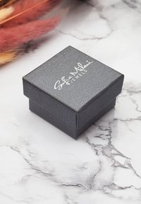 Scatola di gioielli nera con texture, dotata di chiusura magnetica, con marchio bianco in rilievo sul coperchio, posata su una superficie in marmo.