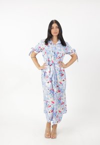 Floral maxi-jurk in lichtblauwe stof, met korte mouwen, een knoopsluiting aan de voorkant en een strikdetail bij de taille. Versierd met kleurrijke bloemenpatronen.