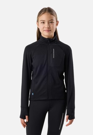 Ung kvinde med glat hår iført en sort jakke med fuld lynlås og matchende bukser, stående mod en ensartet lys baggrund.