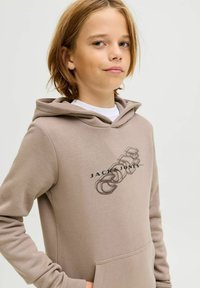 Sudadera color marrón claro con un bolsillo frontal, que presenta un logo gráfico en negro. El material parece suave y liso, con un ajuste relajado y capucha.