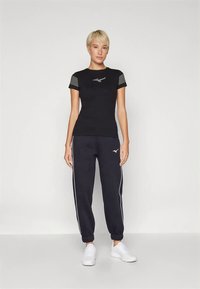 Melna pieguļoša T-krekls ar svītrainām piedurknēm un logotipu krūtīs, kombinēts ar tumši zilām sporta biksēm ar baltām sānu svītrām un logotipu. Baltas sporta kurpes.