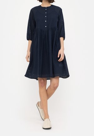Femme portant une robe midi bleu marine perforée avec des manches trois-quarts, des boutons sur le devant et des chaussures blanches sans lacets, se tenant devant un fond blanc.