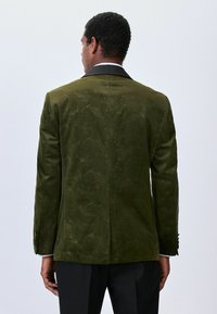 Mees, kes kannab oliivirohelise mustriga sametblazeri, millel on mustad krae, nähtud seljaga lihtsa heleda tausta vastu.