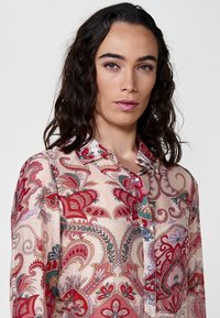 Camicia pulsante in tessuto rosa chiaro, con intricati motivi paisley rossi e verdi, colletto standard e una consistenza morbida e fluida.