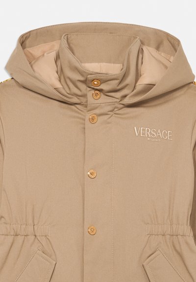 Beige hupparillinen takki, jossa kullanväriset napit, joustava vyötärö, etutaskut ja rintaan brodeerattu "Versace Milano".