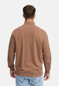 Brown, ribbed long-sleeve pullover with a high collar, soft texture, and cuffed sleeves, worn with light blue jeans.