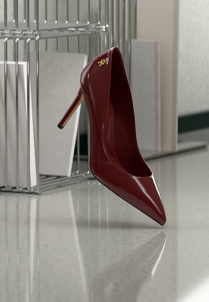 Chaussure à talon haut brillante rouge foncé à bout pointu, posée sur un sol gris réfléchissant près d'un organiseur de dossiers en métal.