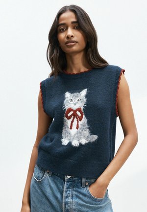 Woman wearing a sleeveless navy sweater with a fluffy cat and red bow design paired with blue jeans, posing against a plain background.