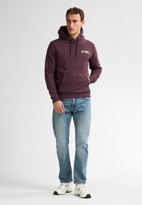 Bordeauxfarbener Hoodie mit Kordelzügen, Fronttasche und Logodetail; getragen mit hellblauen Jeans und weißen Sneakern vor einem neutralen Hintergrund.