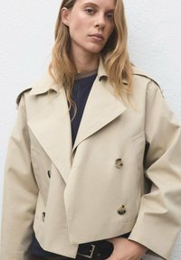 Beige dubbelknäppt jacka med stora slag, mjukt tyg och runda knappar. Bärs över en mörk topp med en kort design.
