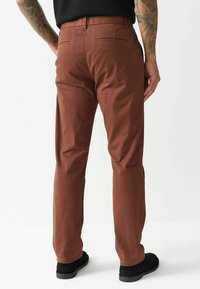 Braune Baumwollchinos im geraden Schnitt, mit zwei Gesäßtaschen, getragen mit schwarzem Schuhwerk. Glatte Textur und maßgeschneidertes Design.