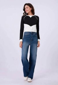 Joven mujer sonriendo, vistiendo una blusa de manga larga en blanco y negro, jeans de pierna ancha azules y zapatillas blancas, de pie contra un fondo simple.