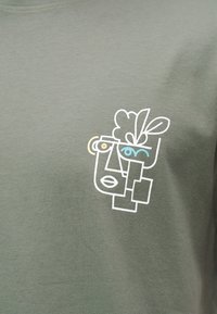 Αφηρημένη γραμμή τέχνης ενός προσώπου με ένα μπλε μάτι και ένα φυτό που μεγαλώνει από την κορυφή, τυπωμένο σε ελιά πράσινο ύφασμα.