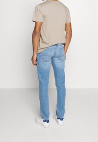 Béžové bavlněné tričko s krátkým rukávem, spárované se světle modrými džíny s kapsami na zadní straně a bílými teniskami s modrými akcenty.