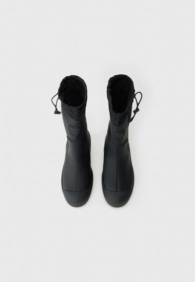 O pereche de cizme negre impermeabile cu mansete ajustabile cu șnur, cu vârf texturat și finisaj mat neted, văzute de sus pe un fundal alb.