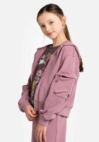 Różowa bluza zip z luźnym krojem, teksturowana tkanina i prążkowane mankiety, nałożona na koszulkę z grafiką z kolorowymi detalami.