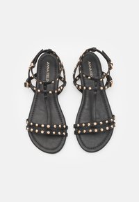 Svarta sandaler med spikade remmar och en platt sula. Utrustad med en spännsrem runt ankeln och en slät, texturerad fotbädd. Metalliska detaljer är närvarande.