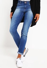 Tajta jeans i en medelblå tvätt, med hög midja, subtil blekning och upprullade mudd. Matchas med vita sneakers.