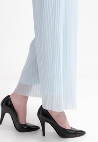 Pieds portant des escarpins noirs à bout pointu, associés à un pantalon large plissé bleu clair transparent, debout sur une surface blanche.