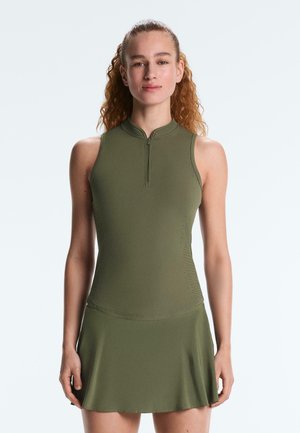 Vrouw met krullend haar draagt een mouwloze, olijfgroene, sportieve jurk met rits en staat tegen een effen lichte achtergrond.