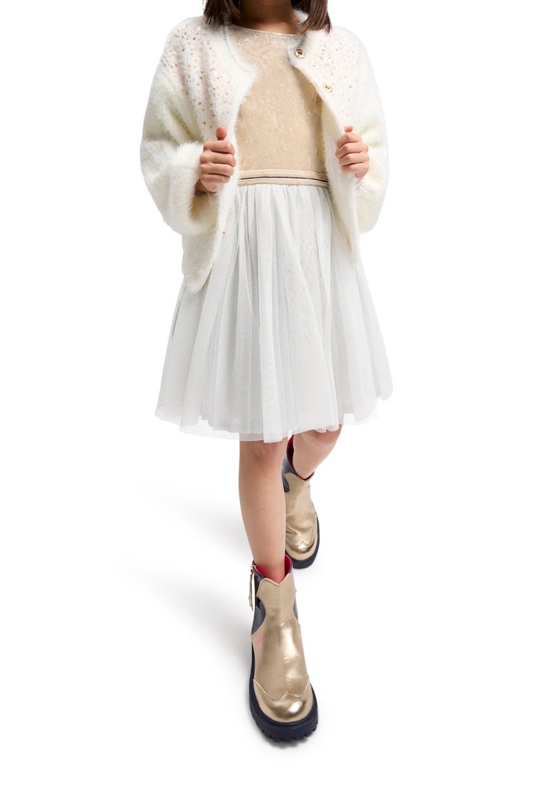 Barn iført hvid tüllnederdel, beige top, hvid blød cardigan med pynt og metallisk guld ankelstøvler, der går fremad.