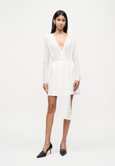 Λευκό φόρεμα τύπου wrap από δαντέλα με βαθύ V-λαιμόκοψη, μακριά μανίκια και μοναδικό ασύμμετρο τελείωμα. Συνδυάζεται με μαύρες γόβες μυτερές.