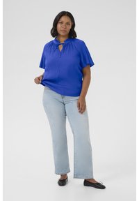 Blouse bleue à manches courtes avec un col haut et un détail à nouer, associée à un jean droit bleu clair et des ballerines noires. Texture douce.
