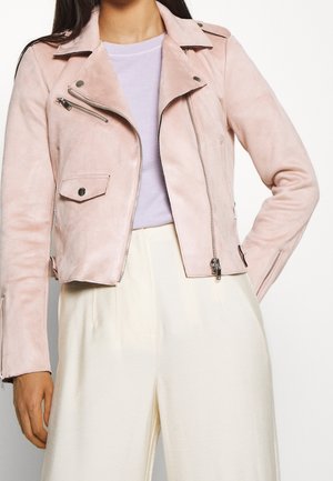 Femme portant une veste de motard en daim rose clair avec des fermetures éclair argentées et un pantalon large taille haute crème sur fond blanc.