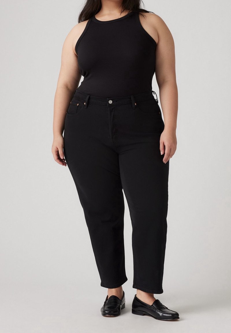 Un débardeur noir à côtelés associé à un jean noir taille haute. Le jean dispose de cinq poches et d'un bouton de fermeture. Des mocassins noirs complètent la tenue.