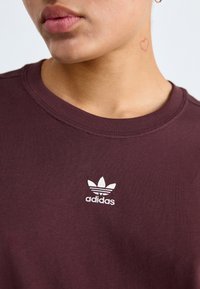 Bordový bavlněný svetr s kulatým výstřihem a krátkými rukávy, se bílým vyšívaným logem Adidas na hrudi. Hladká textura.