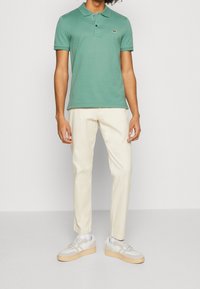 Polo vert avec un col, deux boutons et un petit logo sur la poitrine, associé à un pantalon couleur crème et des baskets claires.