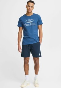 Zils kokvilnas T-krekls ar baltu vintage džinsu rakstu, apvienots ar tumši ziliem šortiem, baltām zeķēm un bēšiem sporta apaviem. Vienkāršs dizains, ikdienišķa piegriezne.