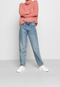 Pull rose texturé à col montant, associé à un jean droit bleu clair et des baskets blanches, posé devant un fond uni.