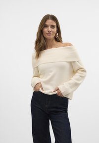 Crème off-shoulder trui gemaakt van zachte stof, met een ribbelige halslijn en lange mouwen, gecombineerd met donkerblauwe denim jeans.
