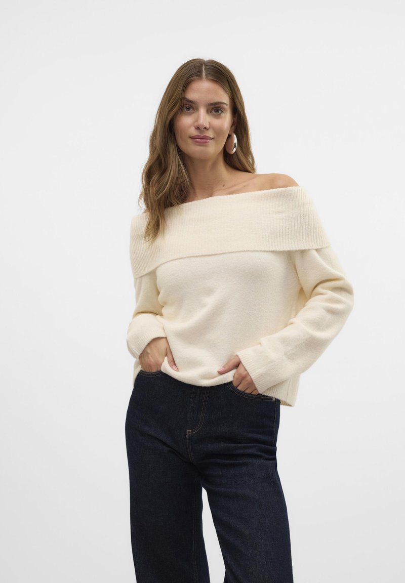 Crème off-shoulder trui gemaakt van zachte stof, met een ribbelige halslijn en lange mouwen, gecombineerd met donkerblauwe denim jeans.