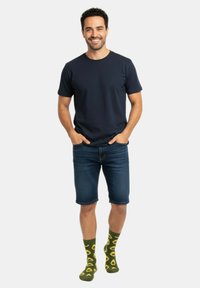 T-shirt a maniche corte blu navy, pantaloni corti in denim e calzini verdi con motivo avocados. Modello in piedi in modo casual, con le mani in tasca, su uno sfondo bianco.