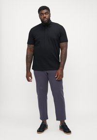 Schwarzes Polohemd mit kurzen Ärmeln, kombiniert mit schmal geschnittenen grauen Hosen. Dunkles Schuhwerk mit strukturiertem Finish rundet das Outfit ab.