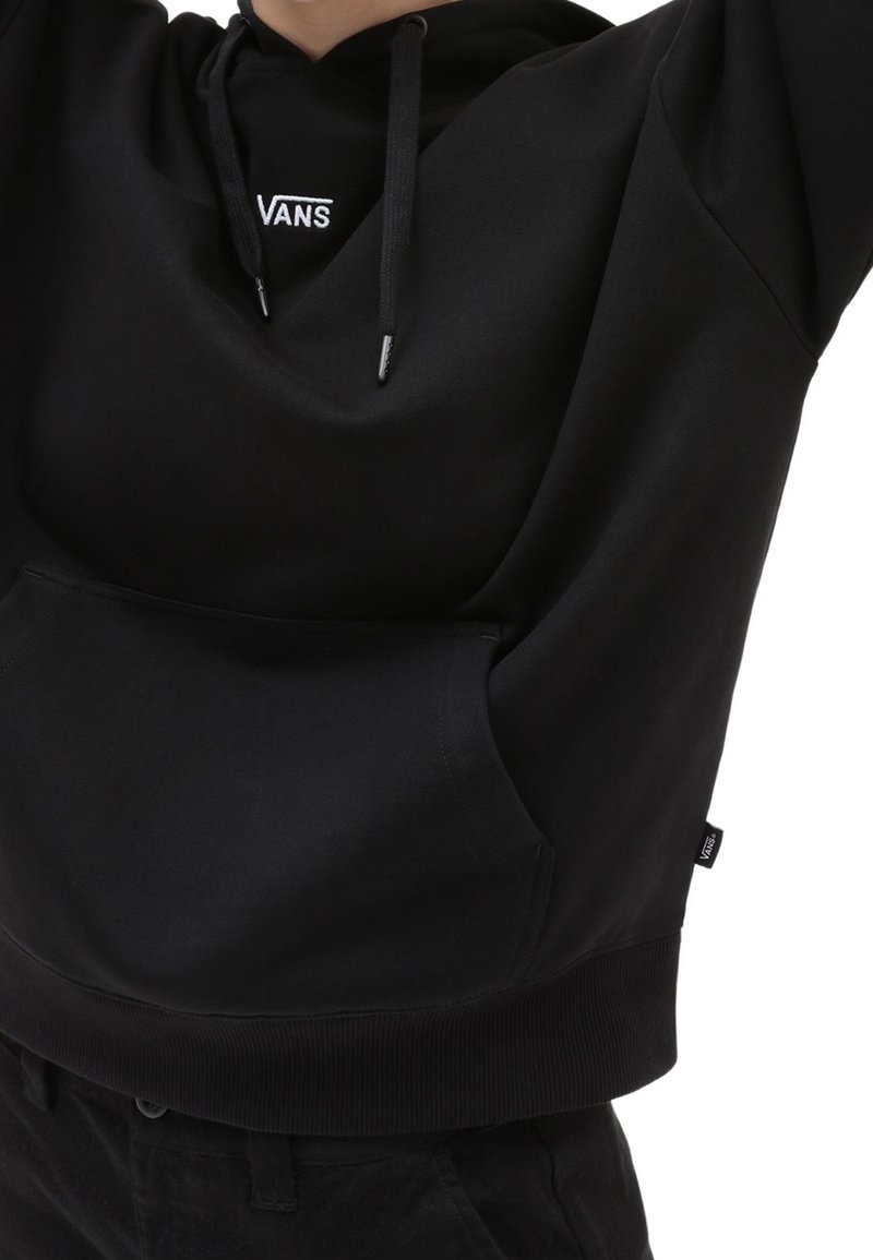Personne portant un sweat à capuche noir Vans avec une poche avant et des cordons, montrant le logo Vans sur la poitrine et l'étiquette latérale.