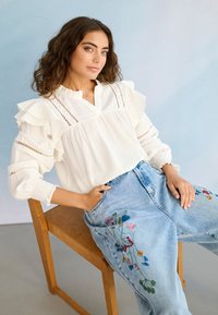 Jeune femme aux cheveux bruns bouclés portant un chemisier blanc à volants et un jean bleu clair brodé, assise sur une chaise en bois.
