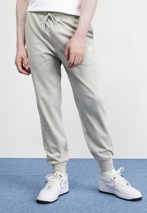 Mann trägt hellgraue Nike-Sweatpants und weiße Nike-Sneaker und steht auf blauem Teppich.