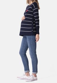 Těhotenský svetr s námořnickými pruhy, červeným srdíčkem, bočními druky a dlouhými rukávy, v kombinaci s modrými úzkými džíny a bílými teniskami.