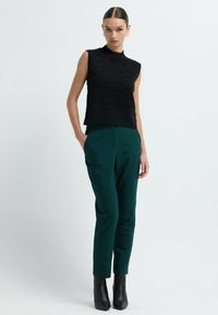 Zwarte mouwloze top met een gestructureerd patroon, gecombineerd met donker groene Tailor-made broeken. Zwarte enkellaarsjes maken de look af.
