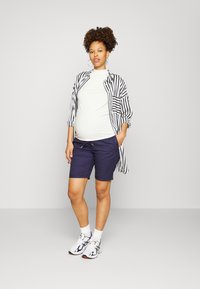 Conjunto de maternidade com uma blusa justa de cor clara, uma camisa oversized listrada em preto e branco, calções azuis-marinho e ténis com padrão.