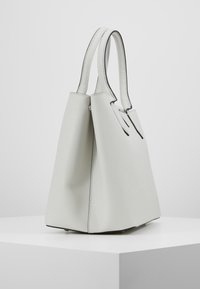Sac fourre-tout gris clair avec une finition texturée, forme angulaire et bord noir. Il dispose de courtes poignées et d'un intérieur spacieux.