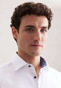 Camicia bianca con colletto a fantasia blu, con bottoni neri e un tessuto strutturato. Il modello indossa la camicia su uno sfondo neutro.
