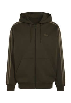 Młody mężczyzna w oliwkowej bluzie z kapturem na zamek marki Adidas z beżowymi paskami na rękawach i beżowych spodniach, stojący na jednolitym tle.