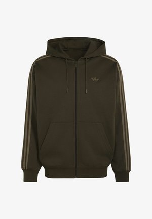 Mladý muž v olivově zelené mikině na zip Adidas s béžovými pruhy na rukávech a béžových kalhotách, stojící před jednobarevným pozadím.