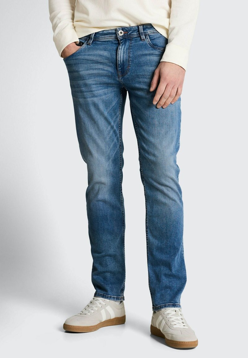 Homme portant un jean slim bleu, des baskets blanches décontractées et une chemise crème à manches longues, debout avec une main dans la poche.